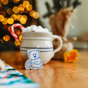 Close up of a wooden bear next to a mug of coco with marshmallows and a candy cane with blurry Christmas lights in the background